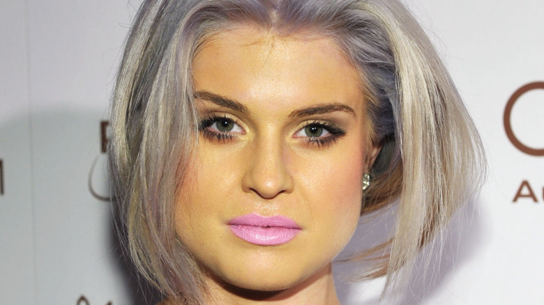 Closeup of Kelly Osbourne on step-and-repeat with gray hair, yellow makeup, and pink lipstick