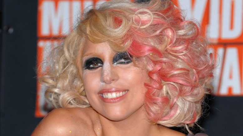 Lady Gaga smiling on step-and-repeat in harsh makeup, pink curls, and white powder on her face