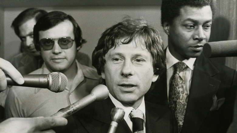 Roman Polanski surrounded by reporters holding microphones
