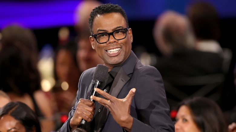 Chris Rock wearing glasses and speaking into a microphone onstage to a crowd of people.