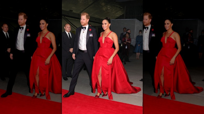 Prince Harry in black tux walking hand in hand with Meghan Markle wearing a red gown at event