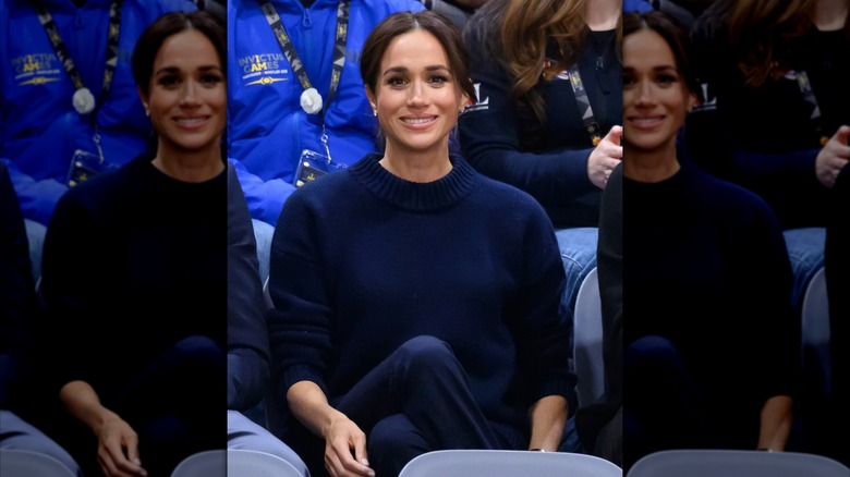 Meghan Markle smiling in bleachers wearing a navy sweater and pants at Invictus Games