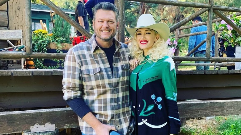 Gwen Stefani wearing a cowboy hat and Blake Shelton wearing a plaid shirt in front of wooden deck