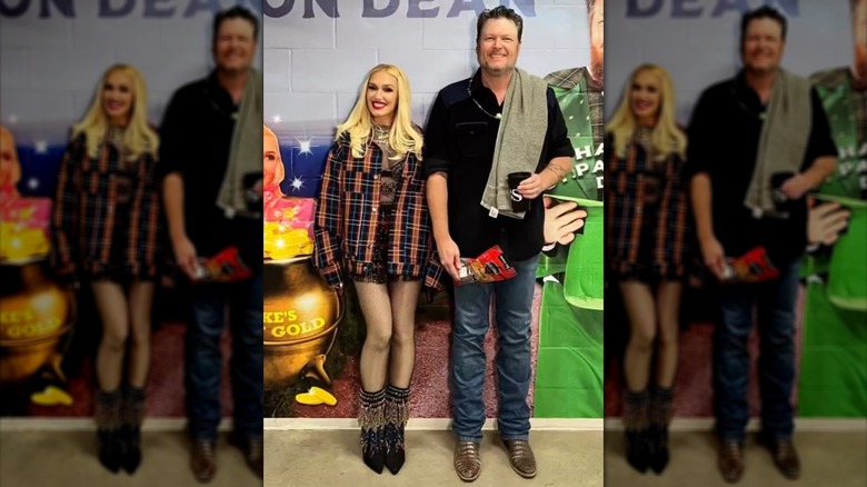 Gwen Stefani wearing a plaid shirt and fishnets while posing next to Blake Shelton, who is holding a package of beef jerky