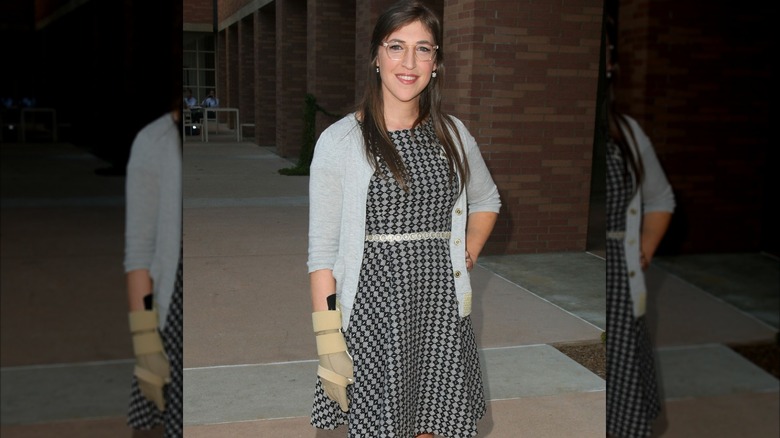 Mayim Bialik wearing checkered dress, gray cardigan, and cast on hand