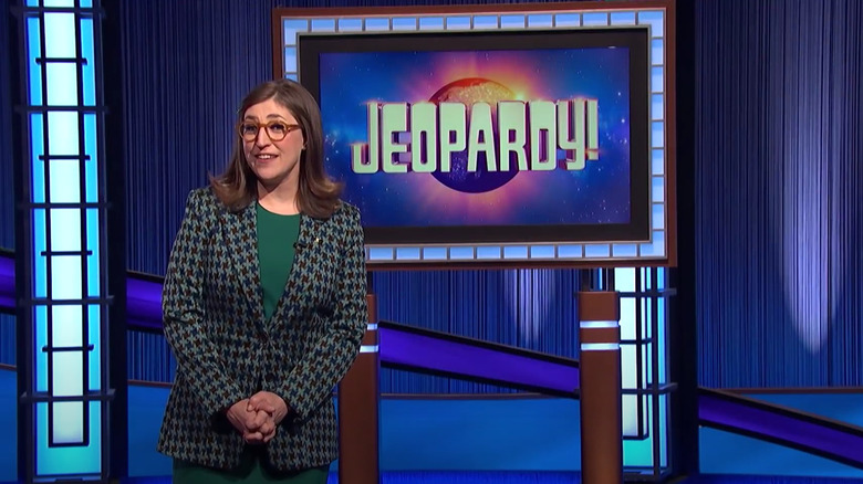 Mayim Bialik wearing green patterned blazer and green dress in front of Jeopardy! screen