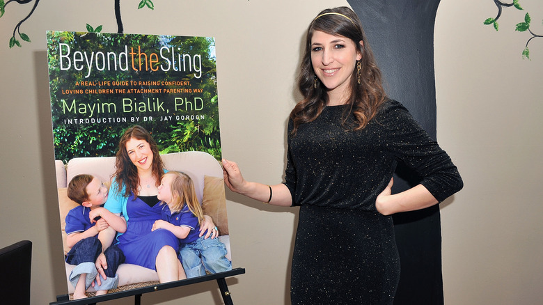 Mayim Bialik wearing a glittery black dress and posing beside a blown-up cover of her book Beyond the Sling
