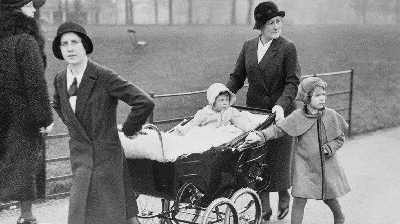 An infant Princess Margaret in a pram and a young Queen Elizabeth II walking alongside two nurses
