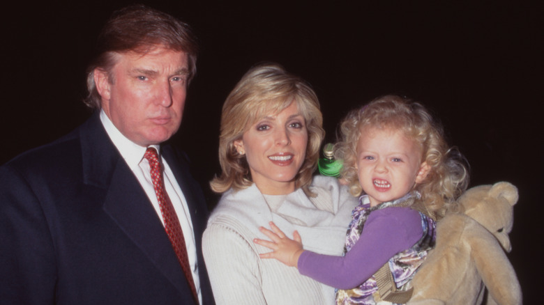 Marla Maples holding a scowling, toddler-aged Tiffany Trump and posing beside Donald Trump