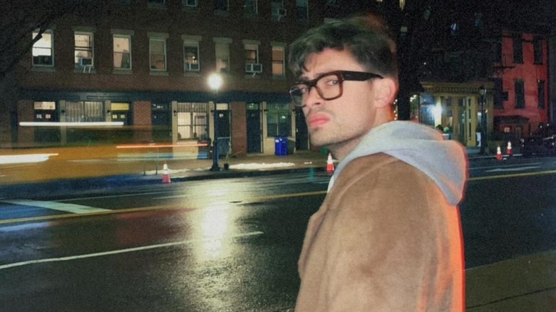 Michael Consuelos standing on a street at night turning toward the camera while wearing glasses and a brown jacket