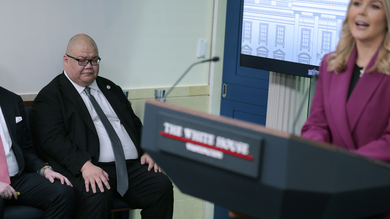 Steven Cheung sits to the side of the lectern and frowns as Karoline Leavitt gives her first news conference