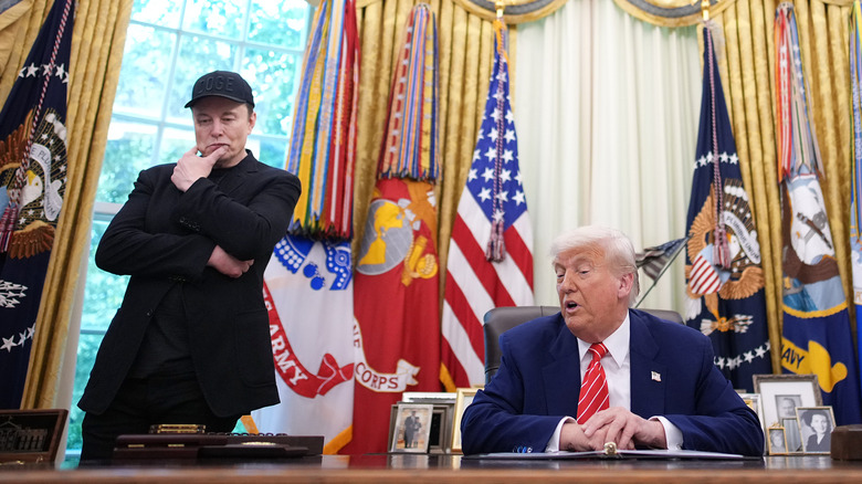 Donald Trump speaks while seated at his desk in the Oval Office as Elon Musk stands beside him looking contemplative