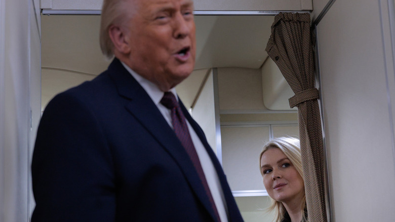 Donald Trump addressing reporters as Karoline Leavitt peeks out from behind a curtain