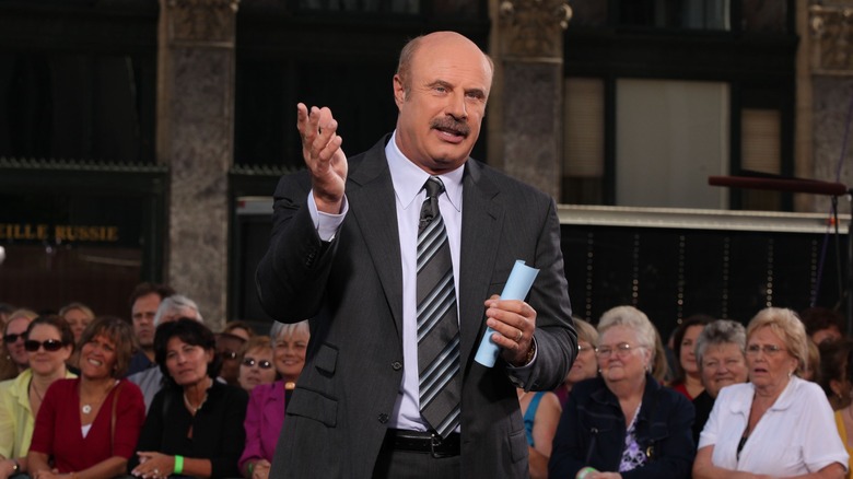Dr. Phil speaking to an audience at an outdoor event while holding a folded piece of paper