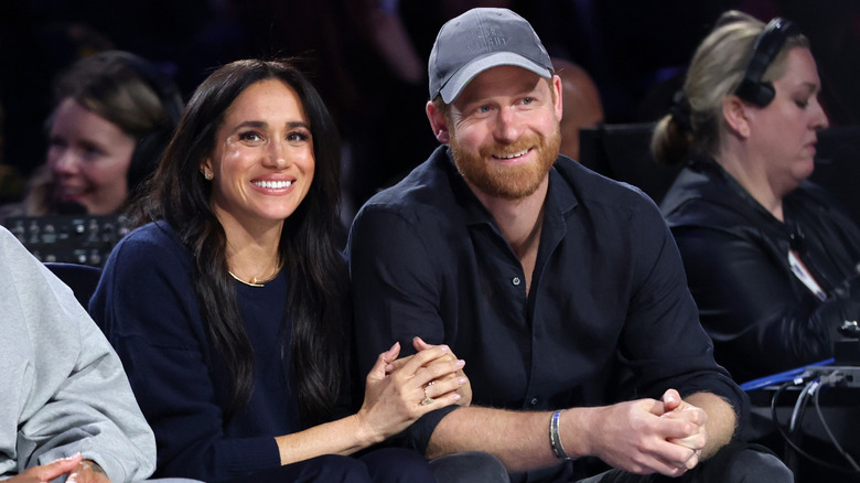 Meghan Markle and Prince Harry linking arms and smiling as they sit in an audience