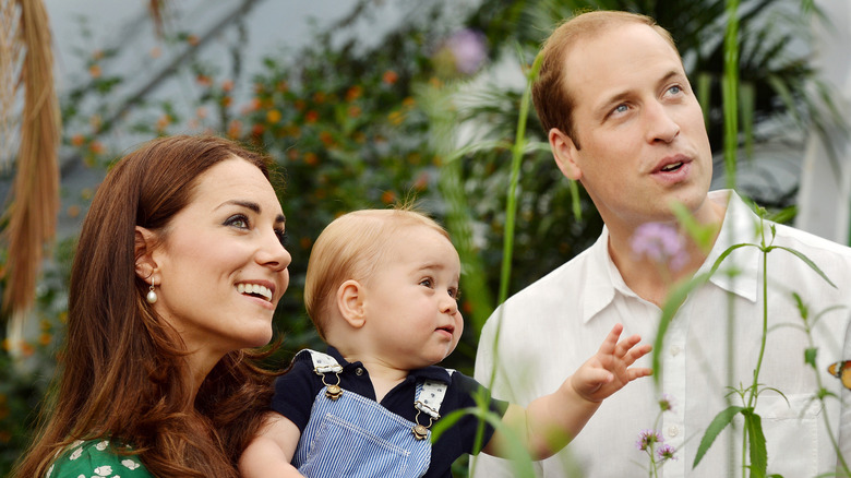 Princess Catherine holding a baby Prince George next to Prince William surrounded by plants and flowers
