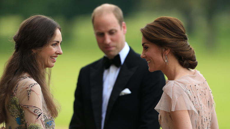 Prince William smiling downward between Rose Hanbury and Princess Catherine who are chatting