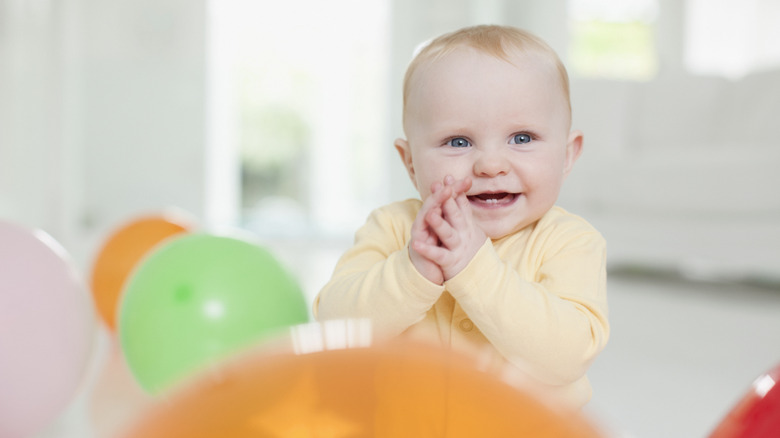 Photo of a smiling baby clasping hands together and surrounded by balloons.
