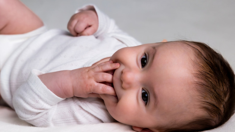 Close-up photo of a smiling baby lying on its back with one hand to its mouth.