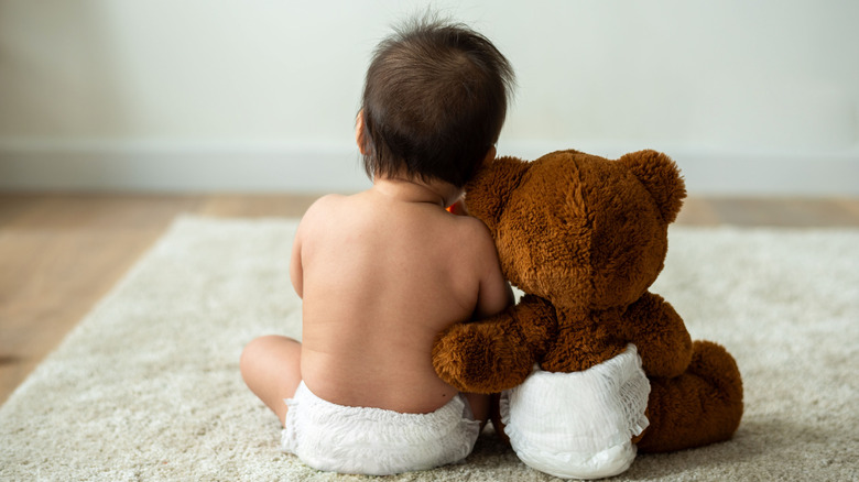 Photo of a baby and teddy bear facing away from the camera and wearing matching diapers.