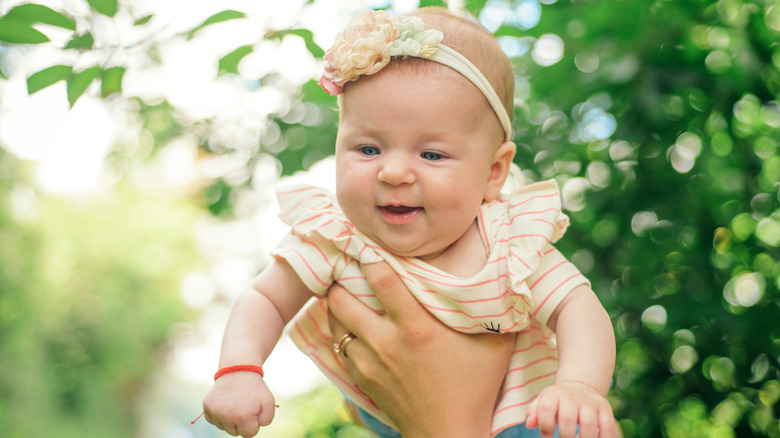 Photo of a smiling baby held up in the air by a parent's hand outdoors.