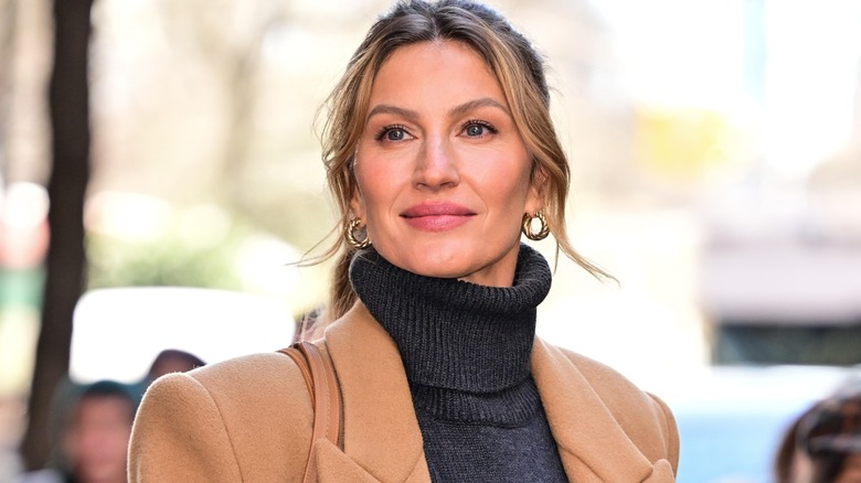 Gisele Bündchen smiling outdoors in a gray turtleneck and camel coat