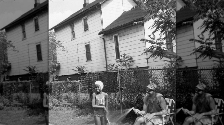 A young Michelle Obama in a swimsuit in a garden while her father, Fraser Robinson, sits in a lawn chair spraying her with water from a hose