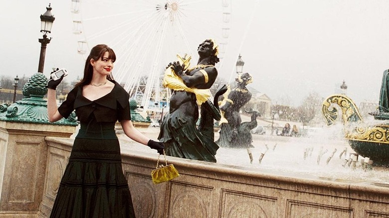 Anne Hathaway as Andy Sachs wearing a black dress and holding her phone up next to a Parisian fountain in "The Devil Wears Prada."
