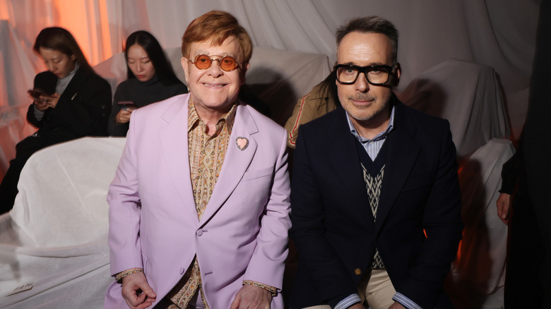 Elton John smiling in a pastel purple suit coat beside David Furnish.