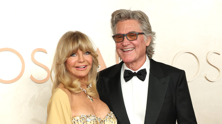 Goldie Hawn smiling in a yellow outfit beside Kurt Russell at the Oscars.