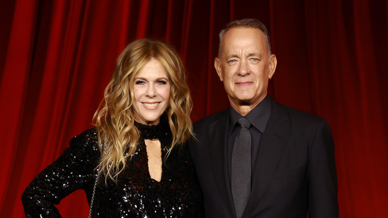 Tom Hanks and Rita Wilson smiling in black formals against red curtains.