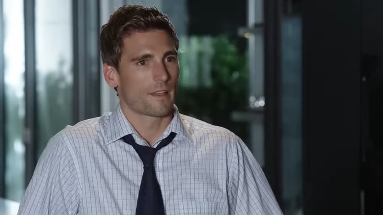 Screenshot of Hallmark Channel movie "A Bride for Christmas," starring Andrew W. Walker. Walker is wearing a button-up shirt and a loose dark blue tie in an office setting and is speaking to somebody off-screen.