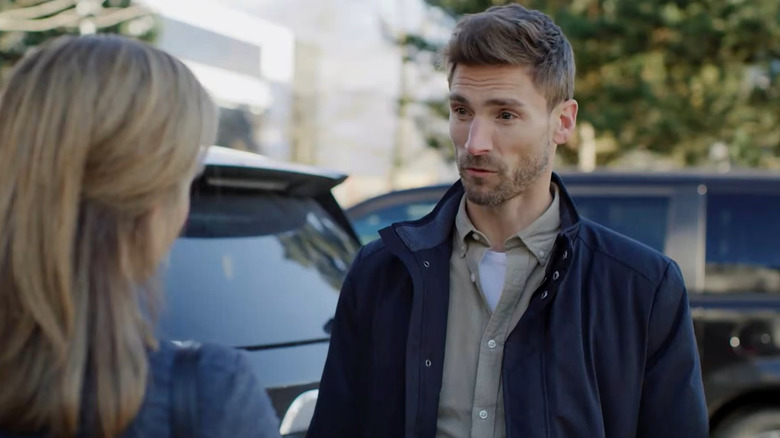 Screenshot of Hallmark Mystery movie "Curious Caterer: Dying for Chocolate," starring Andrew Walker and Nikki DeLoach. Walker is caught in conversation with DeLoach, who is blurred but visible in the foreground, and Walker is wearing a dark blue jacket, beige button-up shirt, and a white T-shirt underneath. There are black cars and trees visible behind him.