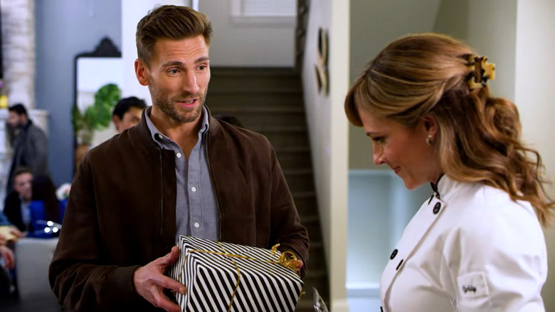 Screenshot of Hallmark Mystery movie "Curious Caterer: Grilling Season," starring Andrew W. Walker and Nikki DeLoach. Walker is holding out a black and white stripe-papered gift box with a gold ribbon to DeLoach, while wearing a brown jacket and blue-grey button-up shirt. DeLoach is wearing a white chef's jacket with her hair half-up/half-down in a tortoiseshell claw clip. Behind the two are other people in a restaurant setting, plus some stairs.