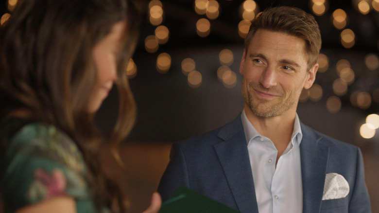 Promotional image for Hallmark movie "Kentucky Roses," starring Andrew W. Walker. Walker is wearing a pale blue shirt and grey-blue jacket with a pocket square, and he's smiling at his co-star, who is blurry in the foreground. Blurred twinkle lights are behind him.