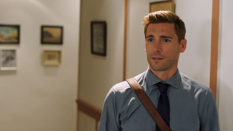 Screenshot of Hallmark original movie "Love In Design," starring Andrew W. Walker. Walker is wearing a blue long-sleeved button-up shirt with a dark blue tie, with a brown crossbody bag strap visible across his chest. He's making a gentle expression at someone beyond the camera, and behind him are paintings/photos hung on office walls.
