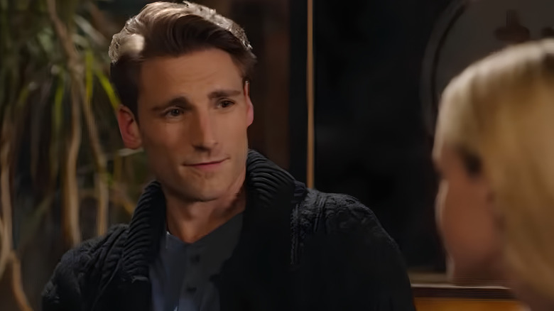 Screenshot of Hallmark movie "Love On Ice," starring Andrew W. Walker. Walker is speaking to a blonde woman in the foreground, looking at her with a soft expression, and wearing a dark blue cable-knit sweater-cardigan and a blue shirt.