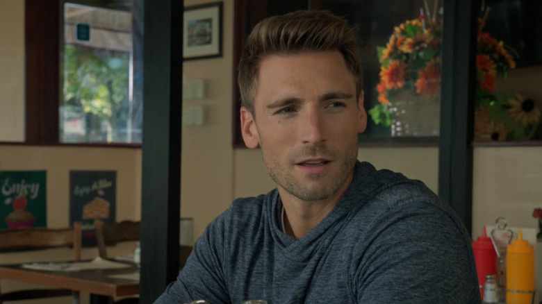 Screenshot of Hallmark movie "Love Struck Cafe," starring Andrew W. Walker. Walker is making a confused/amused expression at someone not seen, as he sits at a cafe table with condiment bottles behind him. Walker is wearing a blue/grey hooded sweater and has visible stubble.