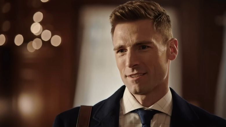 Screenshot of Hallmark movie "Merry & Bright," starring Andrew Walker. Walker is looking at someone not pictured and speaking to them, while wearing a dark blue coat, white grid shirt, and navy blue tie, with the brown strap of a bag on his shoulder. Twinkle lights are blurred behind him.