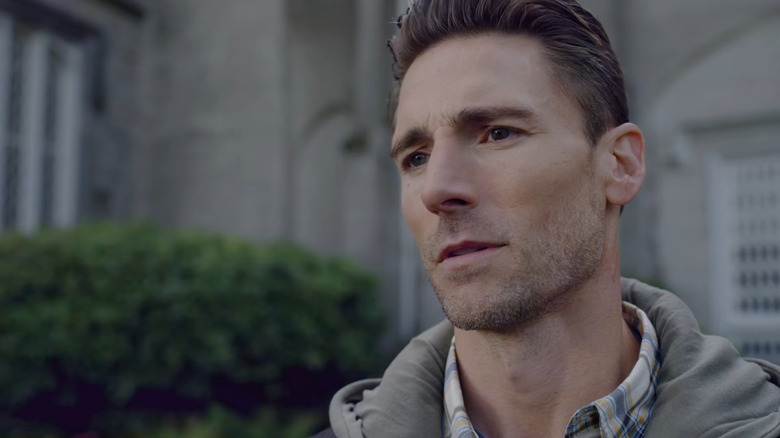 Screenshot of Andrew W. Walker in the Hallmark movie "The Reluctant Royal." Walker is wearing a hooded coat and a plaid shirt, with his hair slicked back, and a grand house and green hedge are visible behind him as he stares at something/someone off-camera.