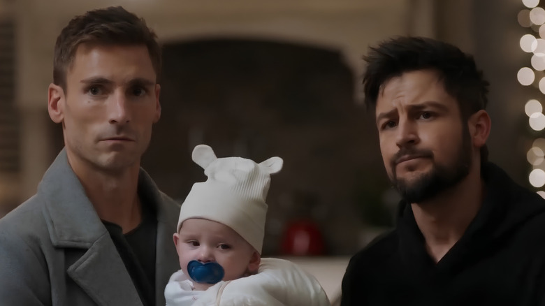 Screenshot of Hallmark movie "Three Wise Men and a Baby," starring Andrew W. Walker and Tyler Hynes. Walker is on the left, Hynes on the right, and the baby is between them. Walker is wearing a grey coat and making an odd expression; the baby is looking away, wearing a white hat with a blue pacifier in his mouth; and Hynes is making a concerned expression at someone past the camera and wearing a black hoodie.