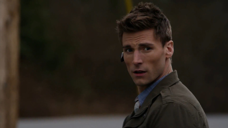Screenshot of Hallmark movie "Wedding Planner Mystery" starring Andrew W. Walker. Walker is making a surprised expression at someone beyond the camera, while wearing a khaki jacket and blue shirt. He also has a Bluetooth headset for a phone in his ear.