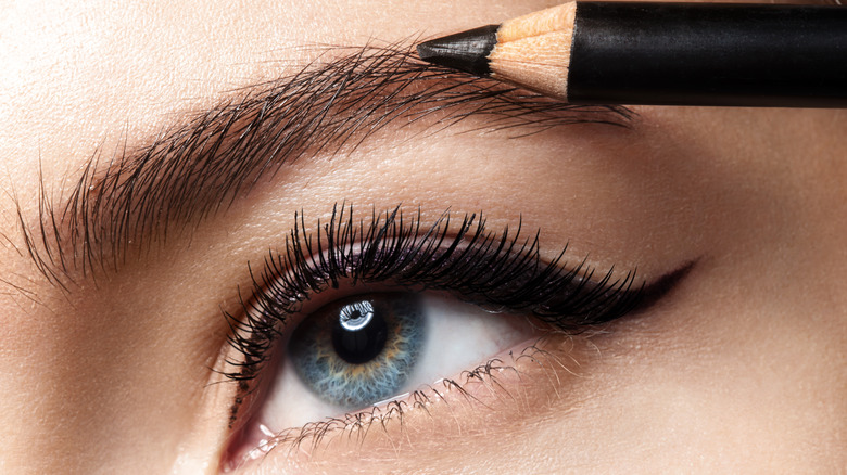 Close up of wooden eyebrow pencil being applied to woman's eye with winged eyeliner