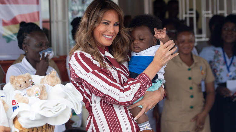 Melania Trump holding an infant while wearing a striped burgundy and white dress