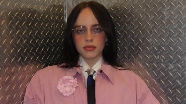 Billie Eilish, wearirng a pink jacket and glasses, standing in front of a metal background.