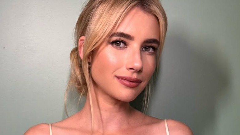 A close up of Emma Roberts standing in front of a sage green wall, smizing.