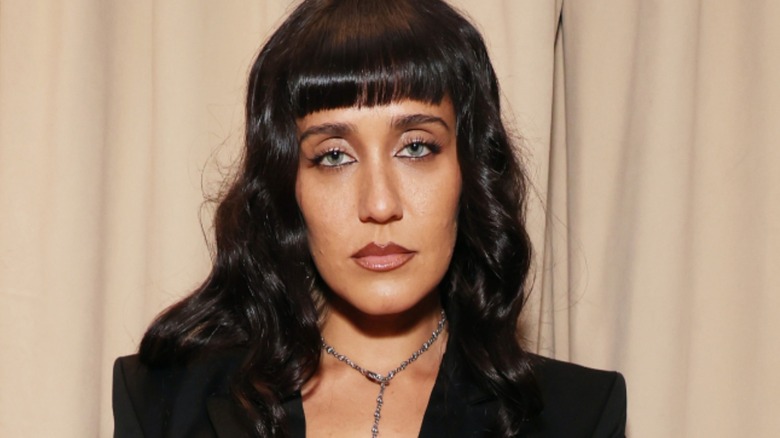 Jesse Jo Stark, in a black suit, standing in front of beige curtains.
