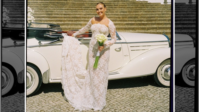 Perrie Edwards leaning on a car in her lace wedding dress, holding a bouquet of flowers