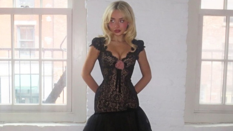 Sabrina Carpenter standing between two windows in a black lace dress with a pink rosette.