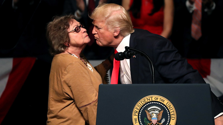 Donald Trump standing behind a lectern with the Presidential seal, squinting, and leaning down to kiss Cuban dissident Cary Roque as she tilts her chin up.
